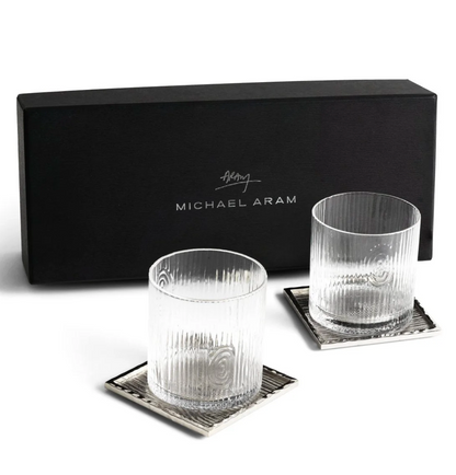 Two ribbed glass tumblers and square silver coasters from the Michael Aram Ivy & Oak Glass and Coaster Gift Set are displayed in front of a black branded box, showcasing rustic elegance.