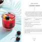 A vibrant red cherry mocktail with ice, topped with a lemon slice and cherry, sits on a table adorned with fresh cherries and lemon wedges. Ideal for fans of delicious non-alcoholic beverages, this enticing drink is featured in Monday Night Mocktails by Simon & Schuster, alongside easy-to-follow mocktail recipes displayed beside the glass.