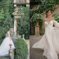 Two women in elegant bridal gowns pose outdoors; one stands on stone steps near lush greenery, while the other lifts her flowing gown from the Monique Lhuillier collection by Penguin Random House as she walks on gravel beside a stone wall adorned with plants.