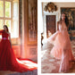 Two women pose indoors amid ornate, classic decor; one stands by a window in a flowing red Monique Lhuillier gown from Penguin Random House, while the other walks through the room wearing a light pink ruffled dress reminiscent of red-carpet styles.