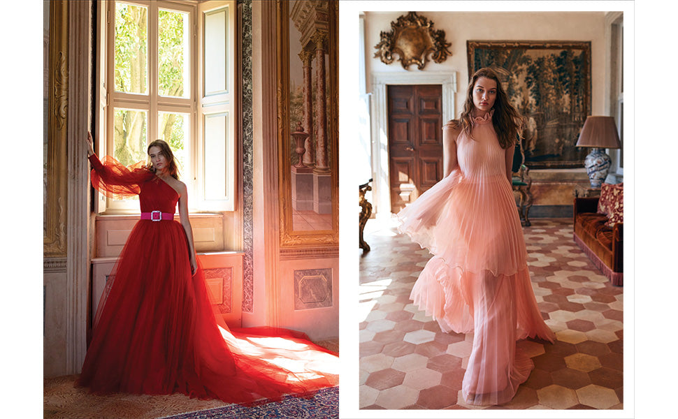 Two women pose indoors amid ornate, classic decor; one stands by a window in a flowing red Monique Lhuillier gown from Penguin Random House, while the other walks through the room wearing a light pink ruffled dress reminiscent of red-carpet styles.
