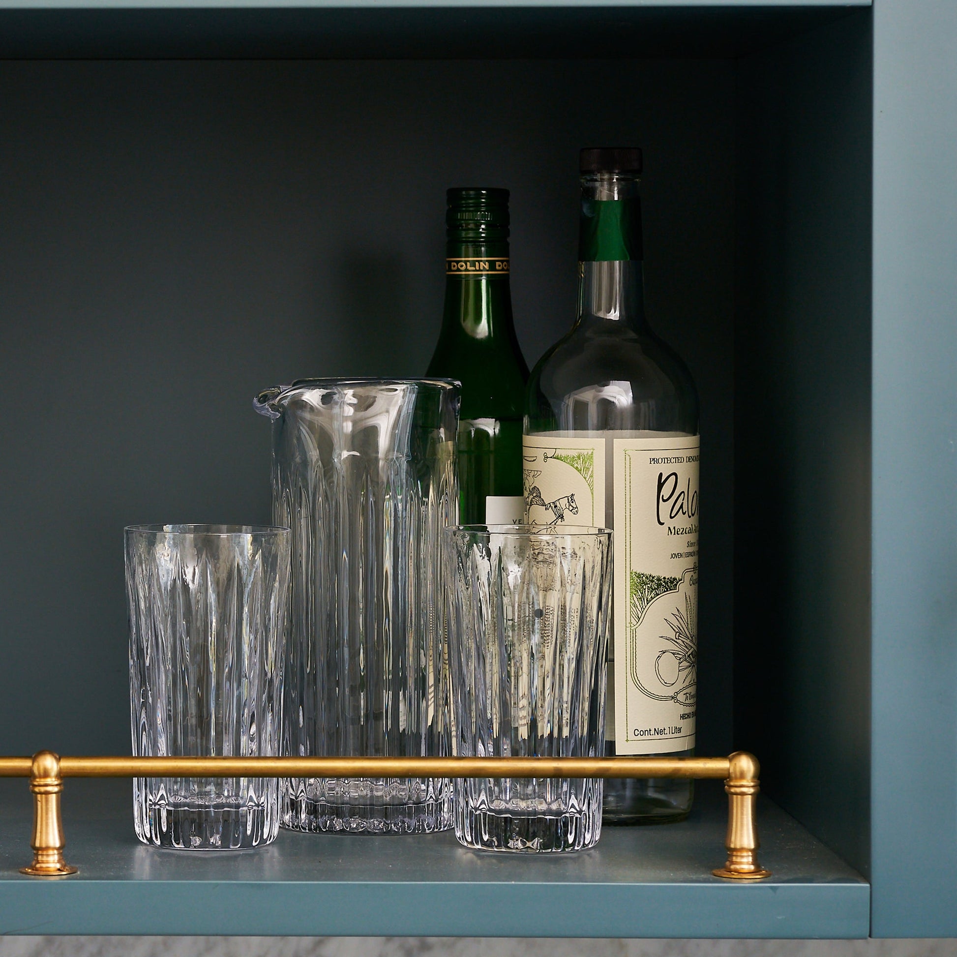 The Simon Pearce Montpelier Highball Gift Set, featuring two bottles, four clear fluted highball glasses, and a pitcher, is arranged on a shelf with a gold rail.
