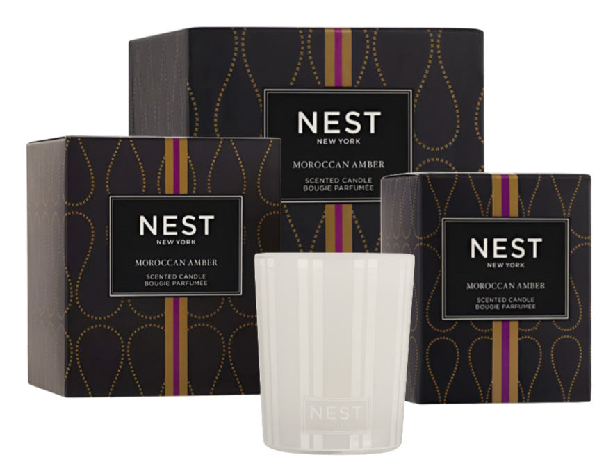 A white Nest Moroccan Amber Candle sits before three black and gold boxes from the Moroccan Amber Candle Collection.
