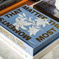 The PrintWorks "Moments That Matter The Most" blue floral coffee table photo album with a cotton cover sits atop two books on a marble surface.