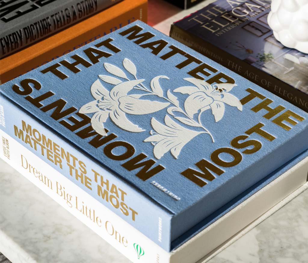 The PrintWorks "Moments That Matter The Most" blue floral coffee table photo album with a cotton cover sits atop two books on a marble surface.
