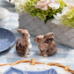 The Juliska Clever Creatures Bunny Salt and Pepper Set features two hand-painted ceramic bunnies with pink ribbons, displayed on a blue and white checkered tablecloth near a white floral centerpiece and blue patterned plates.