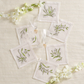The Kim Seybert Vale Cocktail Napkin Set features six white linen napkins with green botanical embroidery, including Lily of the Valley, displayed in a circle next to champagne glasses and white flowers on a cream textured surface.