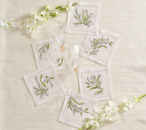 The Kim Seybert Vale Cocktail Napkin Set features six white linen napkins with green botanical embroidery, including Lily of the Valley, displayed in a circle next to champagne glasses and white flowers on a cream textured surface.