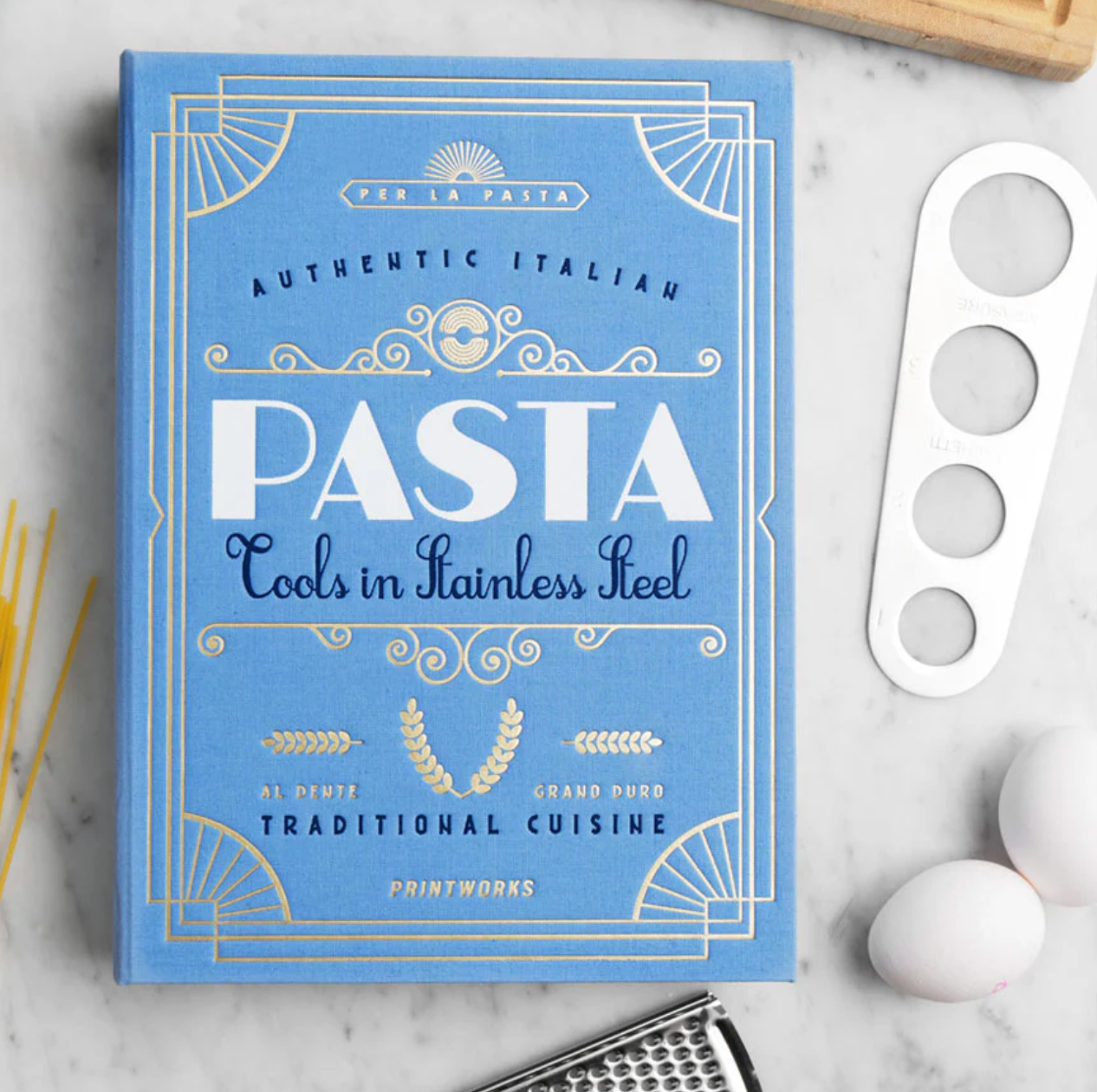 The PrintWorks Pasta Tool Set is displayed on a white surface with pasta, a grater, two eggs, and a blue "Pasta" cookbook—everything you need for memorable Pasta Nights.