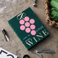 PrintWorks' The Essentials Wine Tools set, featuring a green-covered "Wine" book with a pink grape design, rests on stone surrounded by wine tools, glasses, and a woven basket—making it an ideal gift for any wine enthusiast.