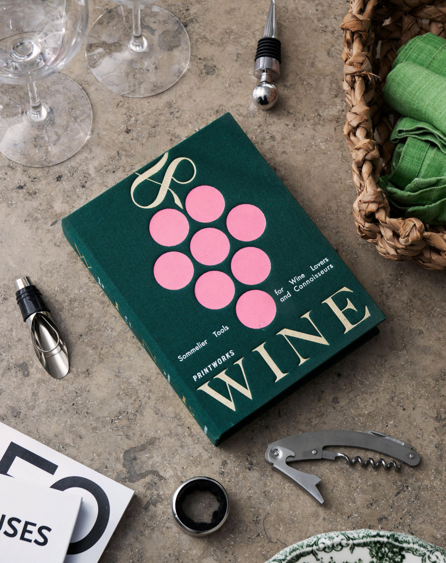 PrintWorks' The Essentials Wine Tools set, featuring a green-covered "Wine" book with a pink grape design, rests on stone surrounded by wine tools, glasses, and a woven basket—making it an ideal gift for any wine enthusiast.