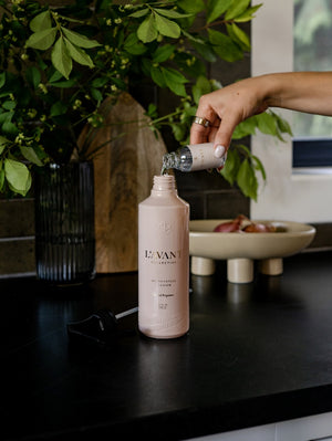 A hand pours Blushed Bergamot Multipurpose Concentrate Refill from a small bottle into a larger L’Avant Collective glass bottle labeled “Multipurpose Surface Cleaner” on a black countertop, with plants and a bowl in the background.
