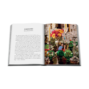 An open book, Boules D'Escalier: The Flávia & Frank Abubakir Collection by Assouline, displays a foreword on the left page and a vibrant image with two people, colorful flowers, green glass decor, and elegant staircase finials on the right.