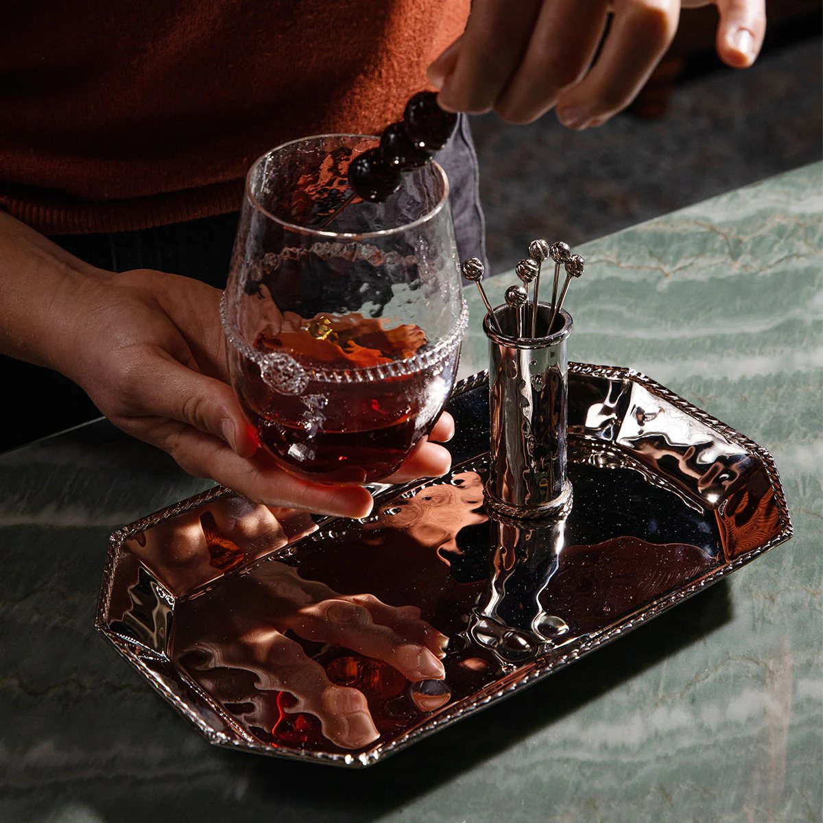 A person places skewered berries into a glass of dark beverage on Juliska's Graham Small Octagonal Tray, with extra skewers in a cup, all elegantly set on green marble as part of a refined barware collection.