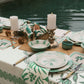 A poolside dining table is set with green and white patterned plates, napkins, candles, and Johanna Ortiz’s Mythic Ecosystem Table Mat Collection.