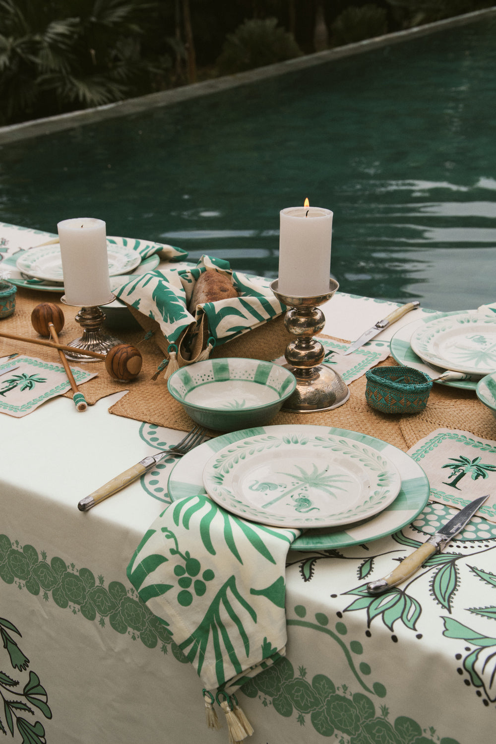 A poolside dining table is set with green and white patterned plates, napkins, candles, and Johanna Ortiz’s Mythic Ecosystem Table Mat Collection.