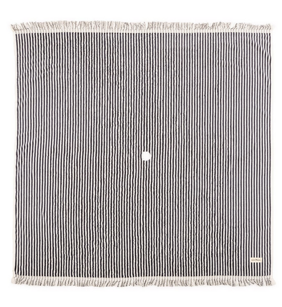 A square black and white striped rug made from 100% cotton, featuring fringed edges and a small white spot in the center from Business & Pleasure The Beach Blanket Collection by Business & Pleasure.