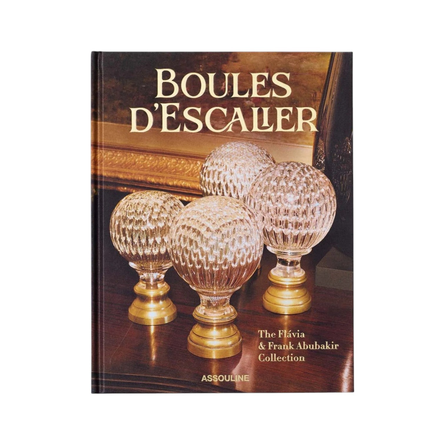 The book cover for "Boules D'Escalier: The Flávia & Frank Abubakir Collection" by Assouline shows four glass spheres with brass bases on wood, inspired by staircase finials from the renowned collection.