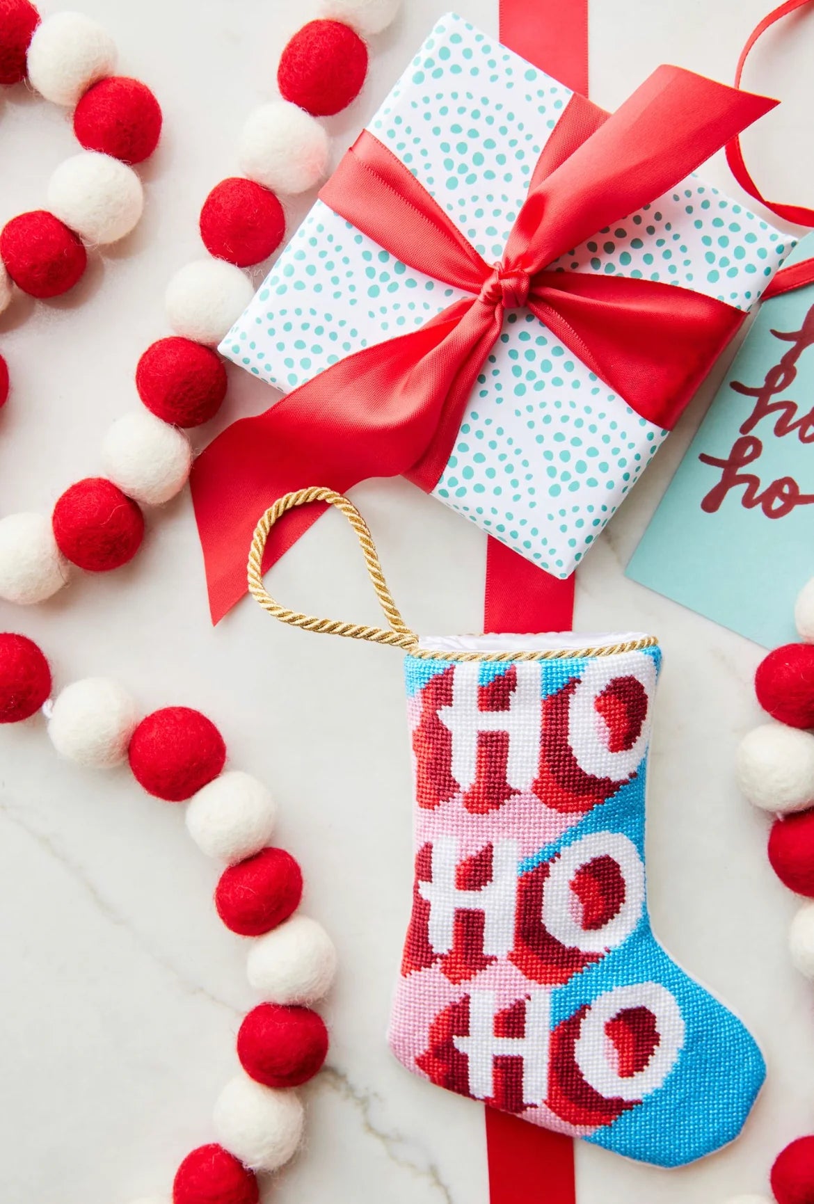 The Bauble Stockings Buru: Ho Ho Ho, Here We Go Needle Point Stocking pairs perfectly with a wrapped gift, tag, and red and white pom-pom garland for festive holiday decor on a white surface.