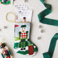 The Bauble Stockings Christmas Birdie Nutcracker Needle Point Stocking holds show tickets and is surrounded by ornaments, a nutcracker, green ribbon, and silver jingle bells on a white surface.