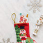 The Bauble Stockings Classic Nutcracker Needle Point Stocking holds a gift card, surrounded by three silver Christmas ornaments and a small nutcracker figurine on a white surface.