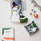 The Frosty Needle Point Stocking by Bauble Stockings, shown with a chocolate bar, assorted charms, jingle bells, and ornaments, makes a charming foundation for your own heirloom Christmas stocking display.