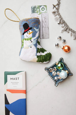 The Frosty Needle Point Stocking by Bauble Stockings, shown with a chocolate bar, assorted charms, jingle bells, and ornaments, makes a charming foundation for your own heirloom Christmas stocking display.