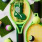 Two yellow tennis rackets, colorful tennis balls, green turf squares, and the Bauble Stockings Tennis Green Needle Point Stocking ornament are arranged on a white surface.