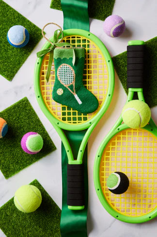 Two yellow tennis rackets, colorful tennis balls, green turf squares, and the Bauble Stockings Tennis Green Needle Point Stocking ornament are arranged on a white surface.