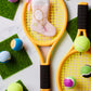 Two yellow toy tennis rackets, colorful toy balls, and green felt squares rest on a white surface. Adding a festive touch is the Bauble Stockings Tennis Pink Needle Point Stocking, perfect for playful holiday decor.