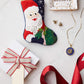 A Bauble Stockings Ho Ho Ho Santa Needle Point Stocking, a "BELIEVE" trinket dish with jewelry, a wrapped gift box, and a red ribbon are displayed on a white surface for a festive holiday scene.