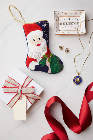 A Bauble Stockings Ho Ho Ho Santa Needle Point Stocking, a "BELIEVE" trinket dish with jewelry, a wrapped gift box, and a red ribbon are displayed on a white surface for a festive holiday scene.