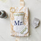 A Bauble Stockings "Mr. Needle Point Stocking" in white and gold marble pattern, displayed with ribbon, wine glasses, wine bottle, and ring box—making a stylish wedding gift or elegant mantle accent.