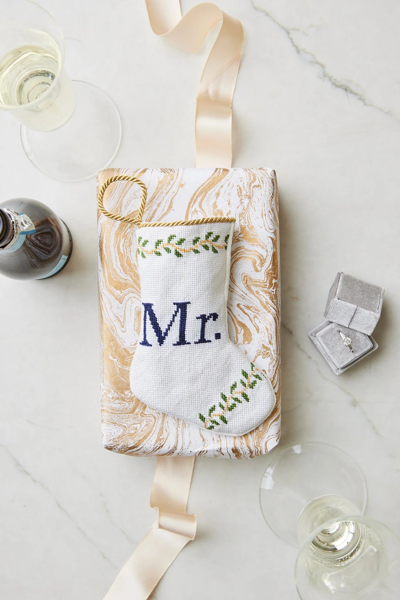 A Bauble Stockings "Mr. Needle Point Stocking" in white and gold marble pattern, displayed with ribbon, wine glasses, wine bottle, and ring box—making a stylish wedding gift or elegant mantle accent.