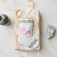 A wrapped gift featuring a hand-stitched Mrs. Needle Point Stocking by Bauble Stockings, a ribbon, two glasses of white wine, a diamond ring box, and a bottle on marble—an ideal bridal keepsake.