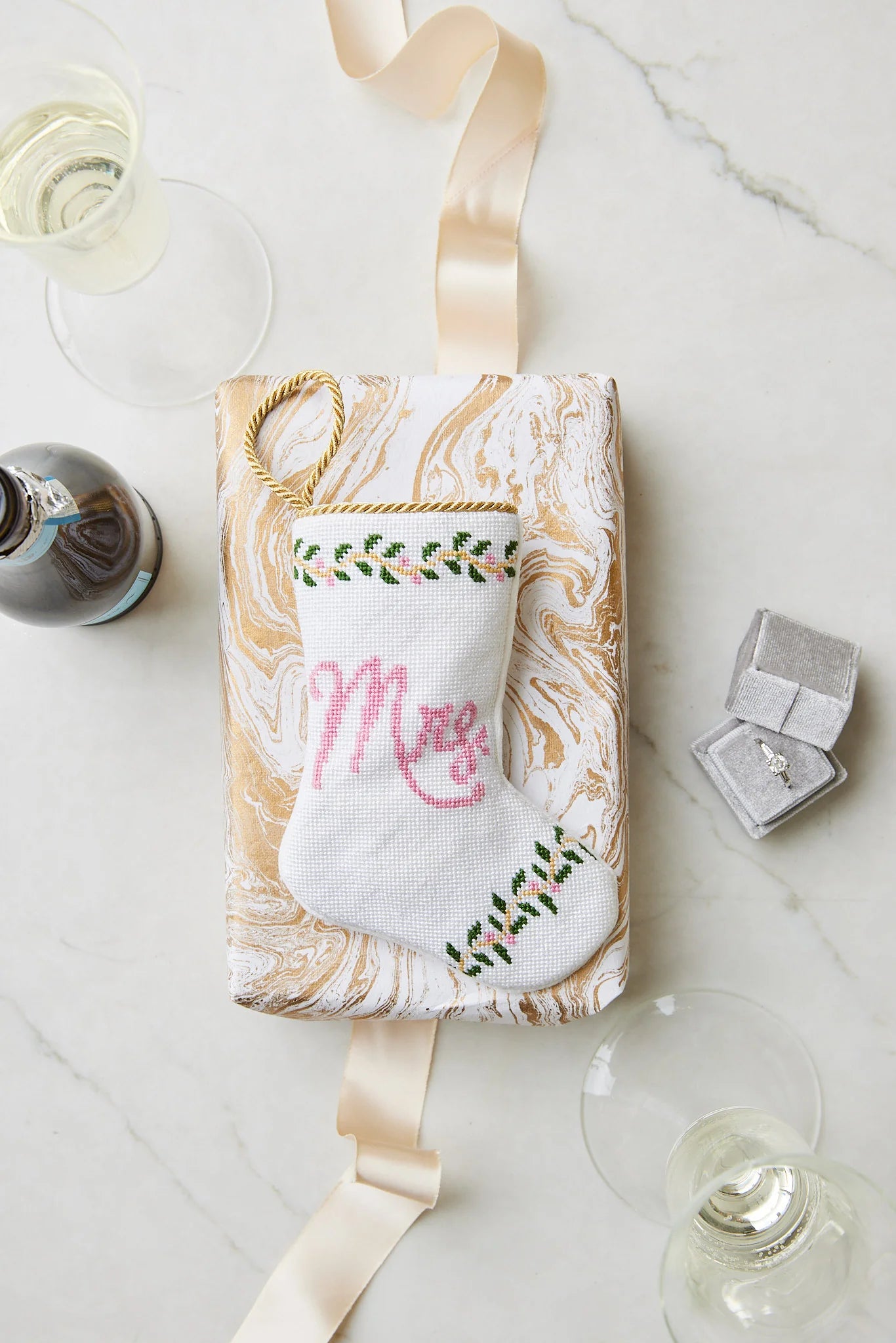 A wrapped gift featuring a hand-stitched Mrs. Needle Point Stocking by Bauble Stockings, a ribbon, two glasses of white wine, a diamond ring box, and a bottle on marble—an ideal bridal keepsake.