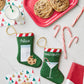 Two green Bauble Stockings "Nice" Needle Point Stockings are on a table with cookies, milk, candy canes, napkins, a red gift, and a red tray—a festive heirloom perfect for your holiday gatherings.