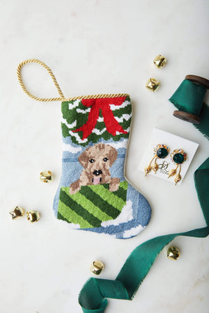 The Bauble Stockings "Open for Joy! Needle Point Stocking" features a hand-stitched puppy design, accented with gold jingle bells, earrings, and green ribbon, displayed on a white surface.
