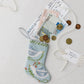 The Bauble Stockings Peace on Earth Needle Point Stocking is displayed on a white surface, filled with international banknotes, coins, airline boarding passes, and topped with a gift tag and string.
