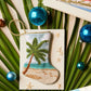The Seaside Serenity Needle Point Stocking by Bauble Stockings features a hand-stitched palm tree design—perfect for adding coastal flair to your holiday decor. Displayed amid palm leaves, blue ornaments, postcards, and starfish.