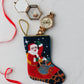 The Bauble Stockings Sleigh Ride Santa hand-stitched needlepoint Christmas stocking holds a gold wristwatch and jewelry box with red ribbon on a white surface, creating an ideal holiday display next to any ornament.