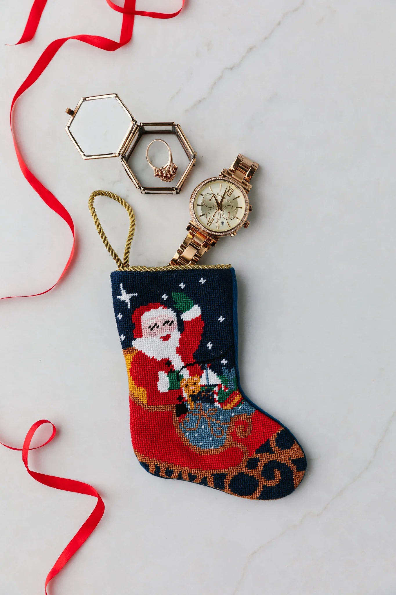 The Bauble Stockings Sleigh Ride Santa hand-stitched needlepoint Christmas stocking holds a gold wristwatch and jewelry box with red ribbon on a white surface, creating an ideal holiday display next to any ornament.