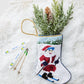 Bauble Stockings’ Slopeside Santa Skiing Needle Point Stocking features a hand-stitched skiing Santa, filled with pine branches and pinecones—charming ski holiday decor with snowflake accents and miniature skis.
