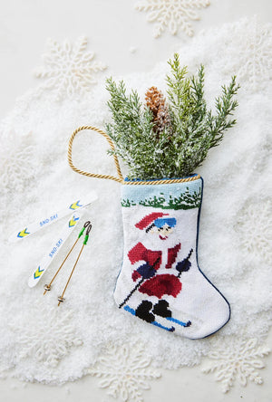 Bauble Stockings’ Slopeside Santa Skiing Needle Point Stocking features a hand-stitched skiing Santa, filled with pine branches and pinecones—charming ski holiday decor with snowflake accents and miniature skis.