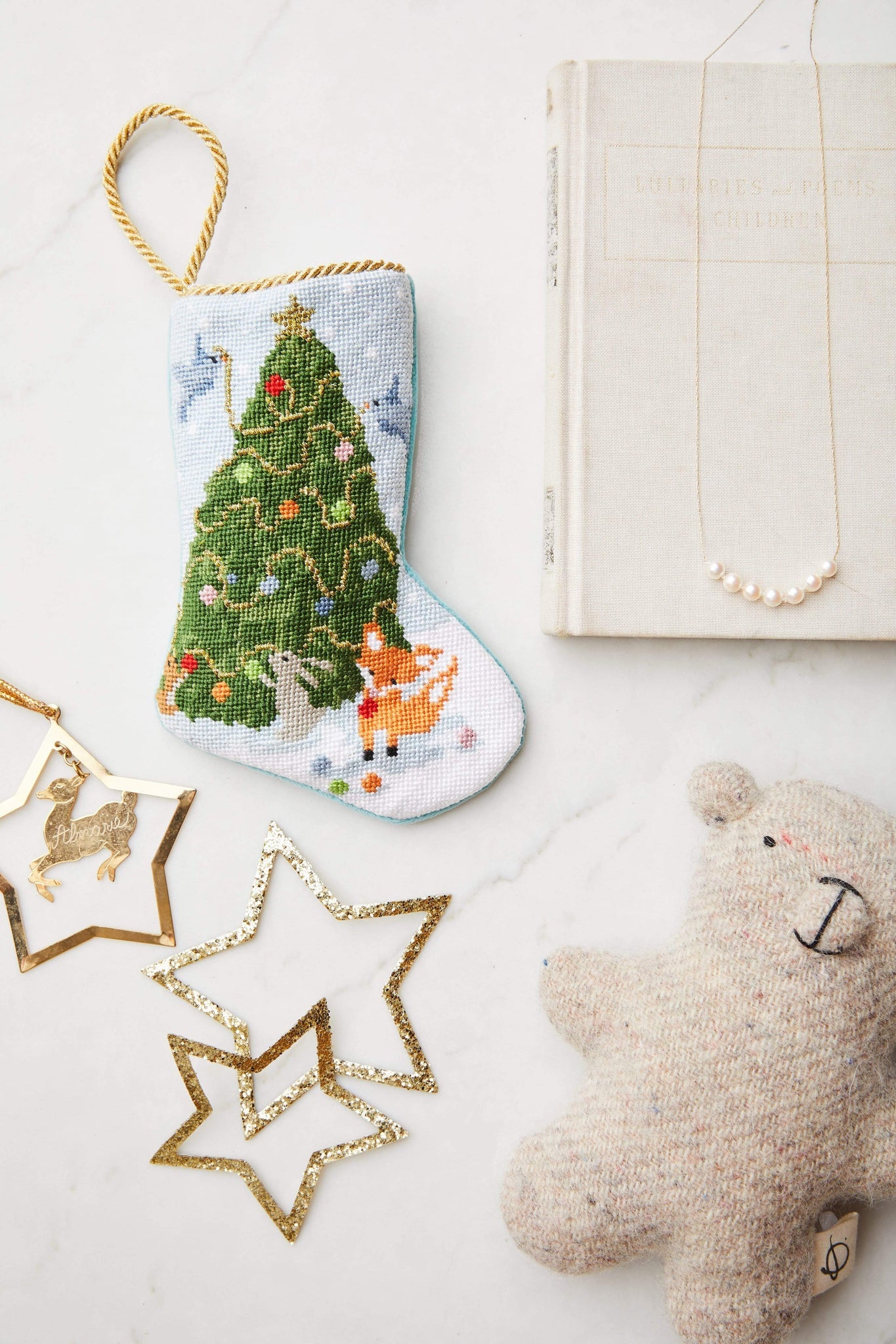 The Woodland Creatures Needle Point Stocking by Bauble Stockings, paired with gold star ornaments, a plush bear, a pearl necklace, and a closed book, creates a charming and festive holiday display on any white surface.
