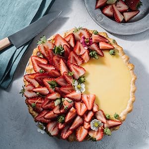 A tart with half of its surface covered in sliced strawberries and the other half displaying yellow custard, garnished with small flowers, sits on a light surface beside a knife and a plate of strawberries—an inspiring scene reminiscent of *The New Pie* by Penguin Random House, a cookbook brimming with innovative pie recipes and expert baking techniques.