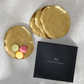Four gold Michael Aram Botanical Leaf Tidbit plates and three pastel macarons are presented in a sleek black branded box on a light surface, exemplifying artisanal luxury.