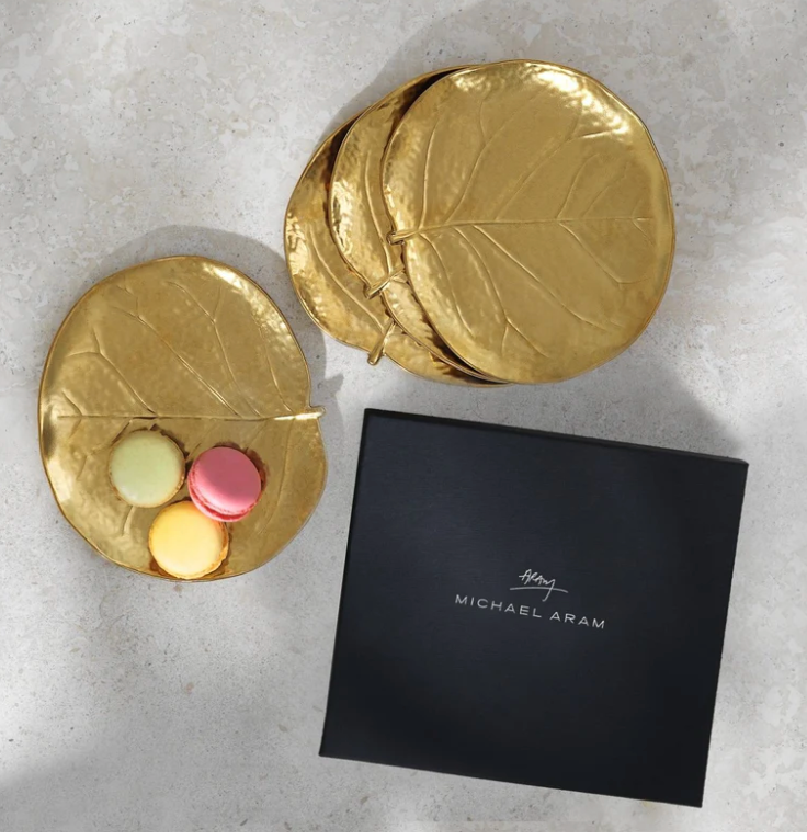 Four gold Michael Aram Botanical Leaf Tidbit plates and three pastel macarons are presented in a sleek black branded box on a light surface, exemplifying artisanal luxury.