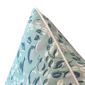 Close-up of the Cowrie Shell Aqua Outdoor Pillow by Associated Design, featuring a white piped edge and abstract leaf shapes in white, blue, and gray on light blue-green, crafted from premium materials.