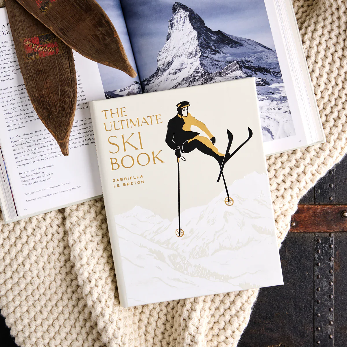 The Ultimate Ski Book by Graphic Image lies on a knitted blanket beside an open magazine with a snowy mountain scene, inviting readers to discover skiing history and culture.